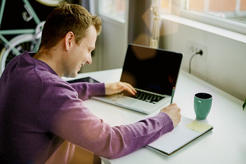 smiling man working on laptop and writing on a notepad